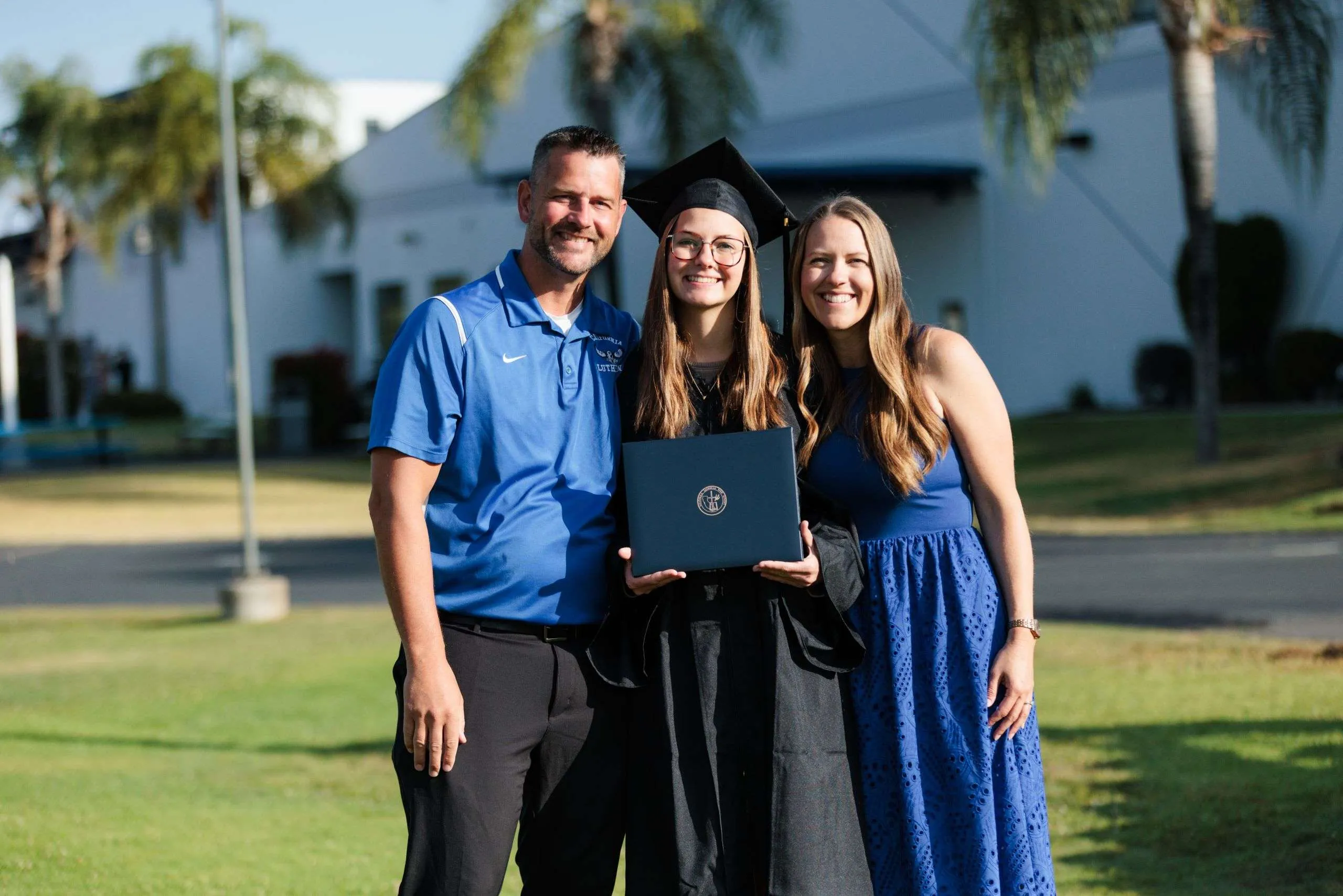 Graduate With Family A graduate in cap and gown holds her diploma between a man in a blue polo and a woman in a blue dress on the campus lawn